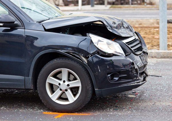 A black car with a damaged front end is parked on the side of the road.