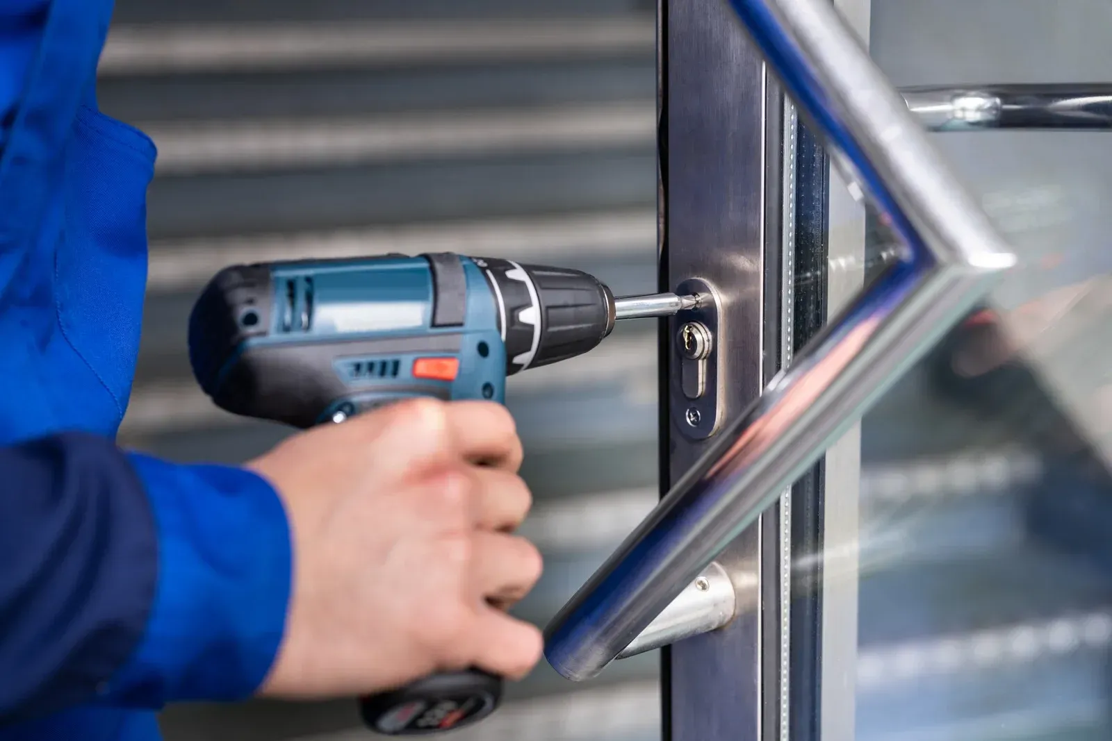 A person in a blue uniform uses a handheld power drill to secure a metal lock mechanism on a door frame.