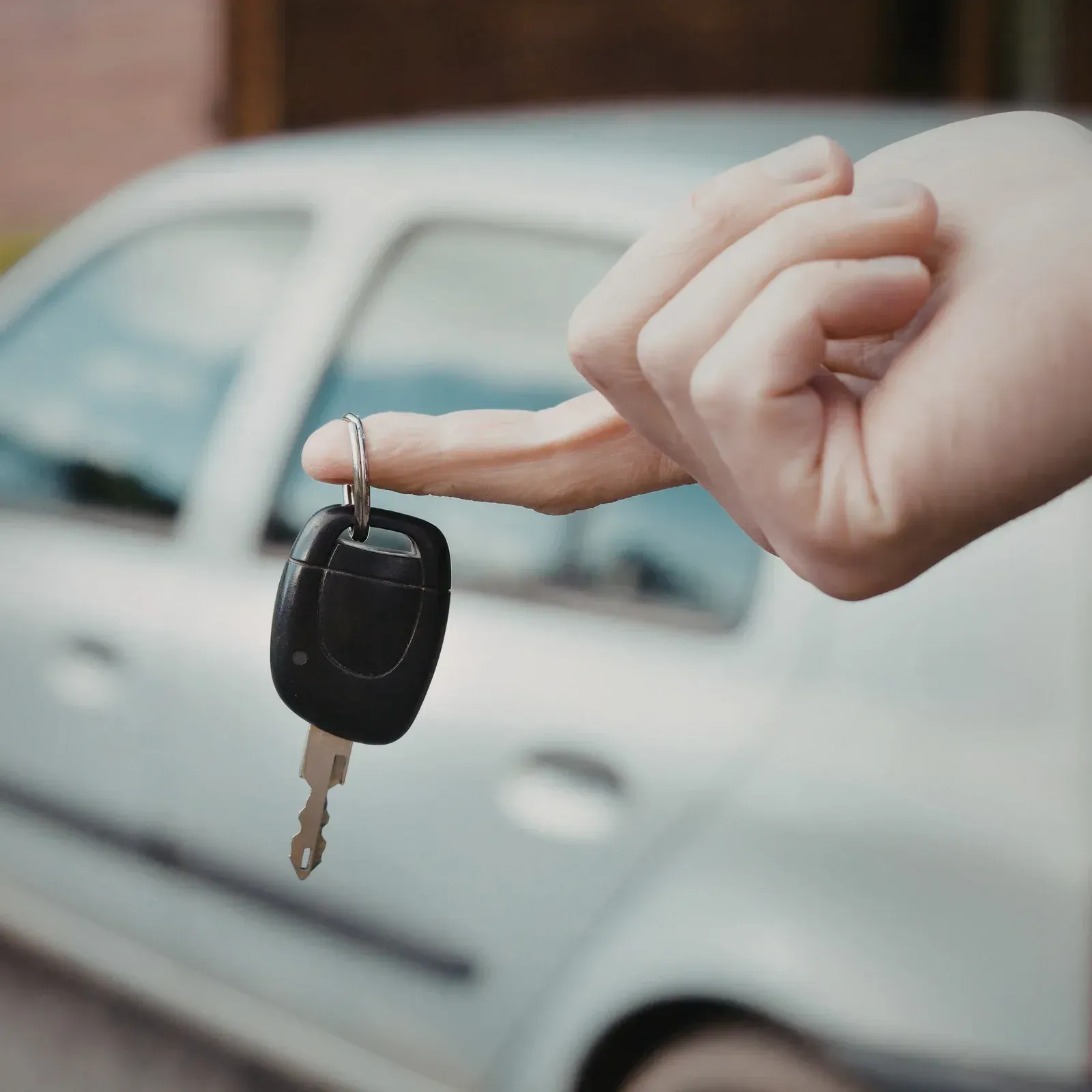 A hand holds a car key by a ring in front of a blurred silver car.