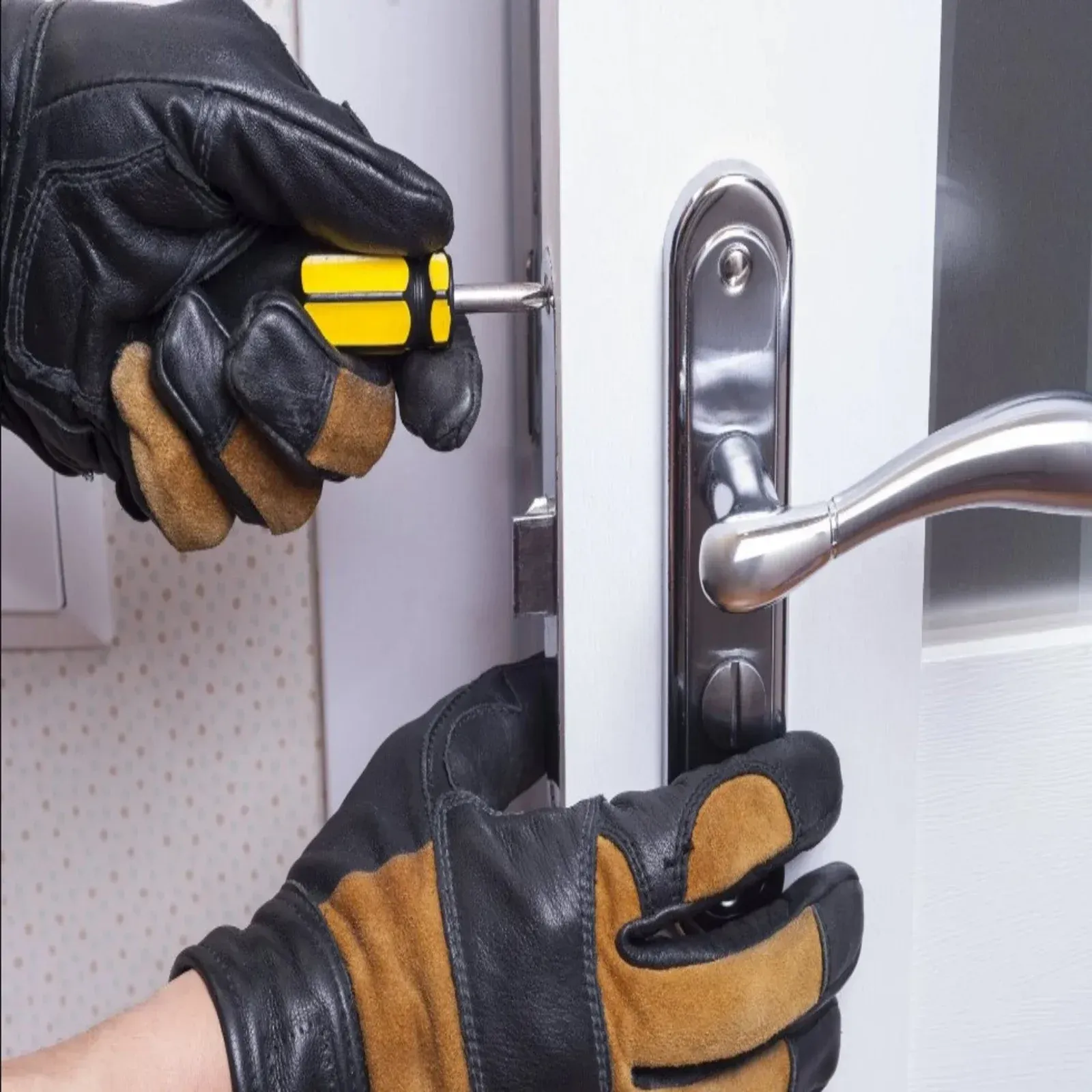 Hands in work gloves use a yellow-handled screwdriver to install or adjust a metal door handle mechanism on a white door.