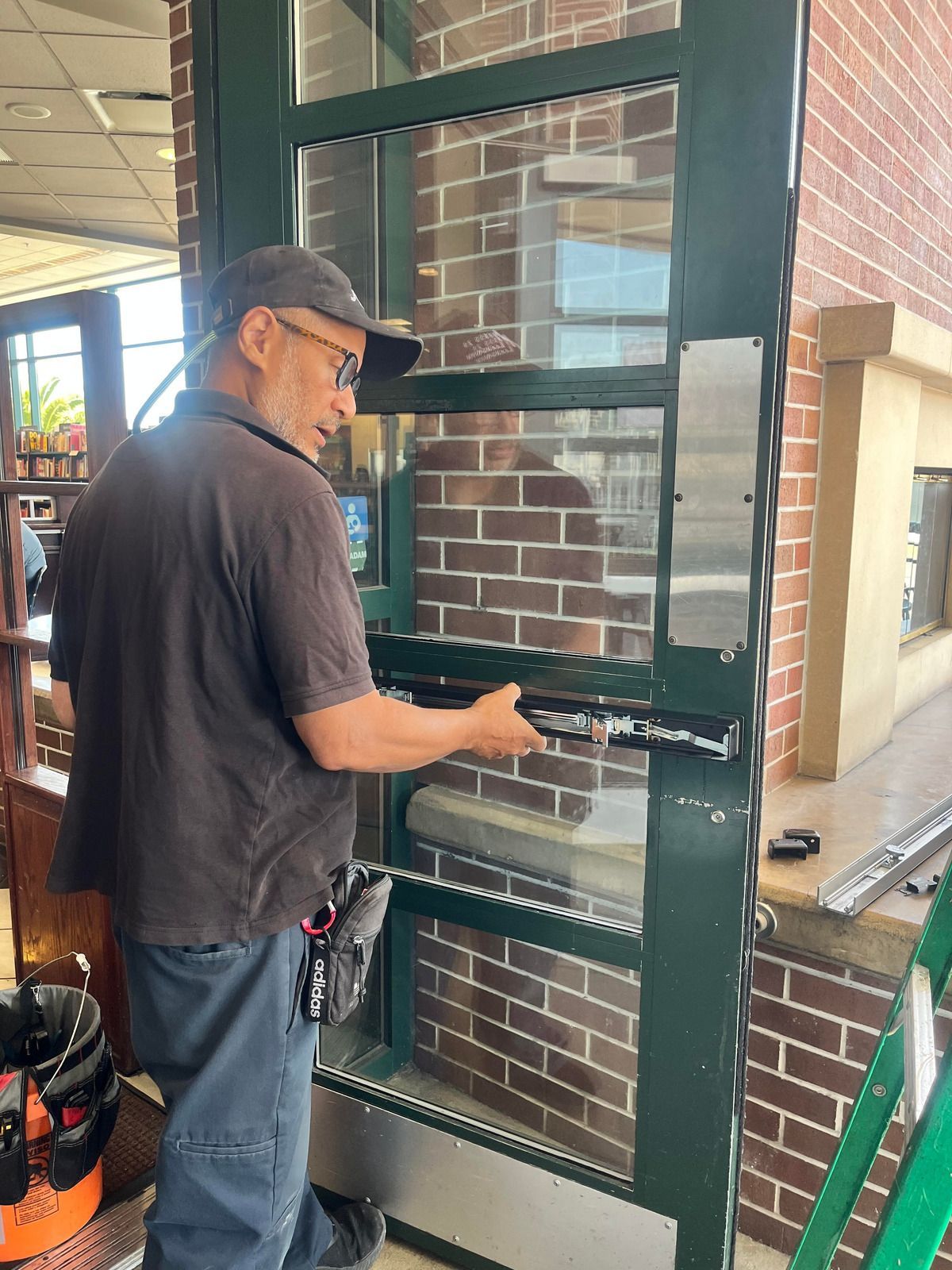 A technician in a dark shirt and cap works on installing a horizontal metal panic bar on a glass commercial door.