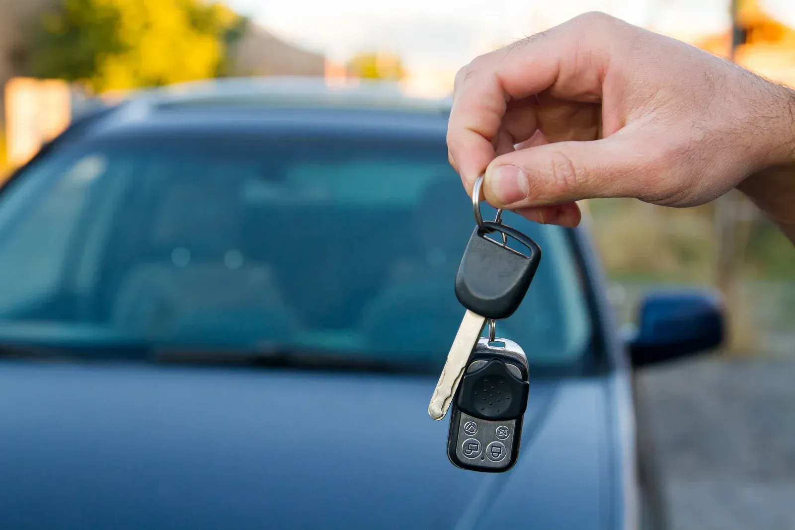 A hand holds a car key and a remote fob in front of a blurred dark blue car.