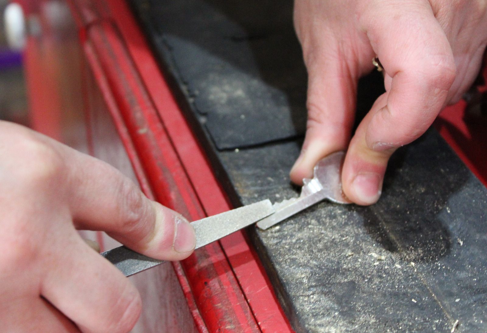 Hands use a metal file to sharpen or smooth the bitting of a silver key on a workshop bench.