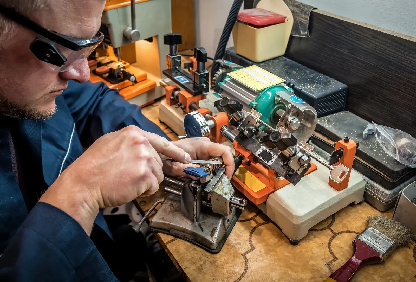 A person wearing safety glasses uses a metal file to work on a key at a workbench equipped with a key cutting machine.
