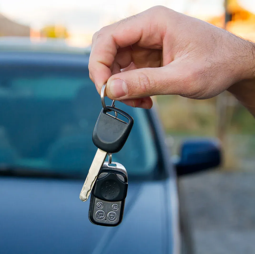 A hand holding a car key and a remote fob in front of a blurred vehicle.