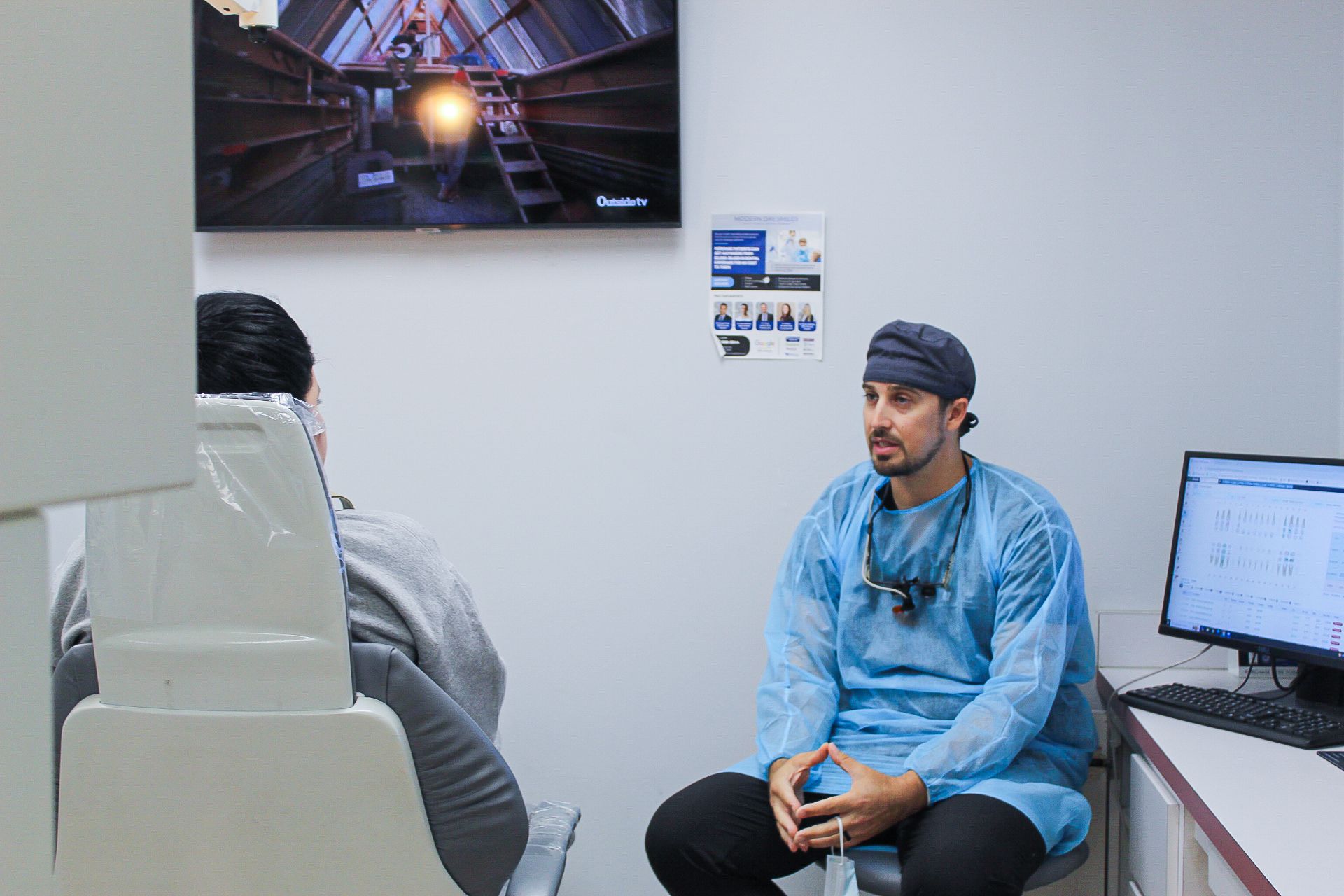 Two people in a clinic office, one seated in blue scrubs beside an exam chair and computer.