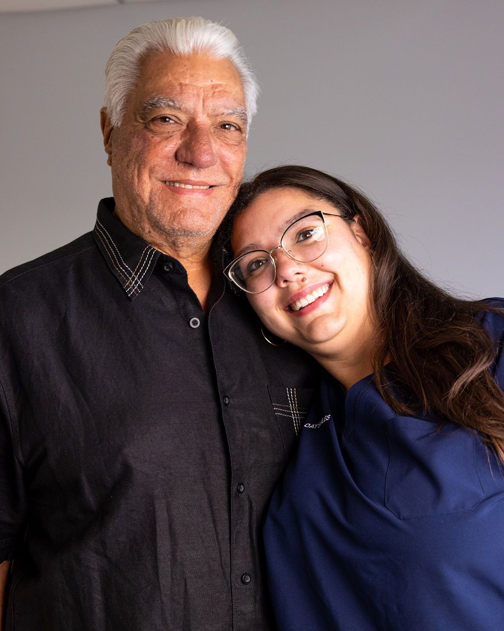 Two people smiling and leaning together against a plain gray background.