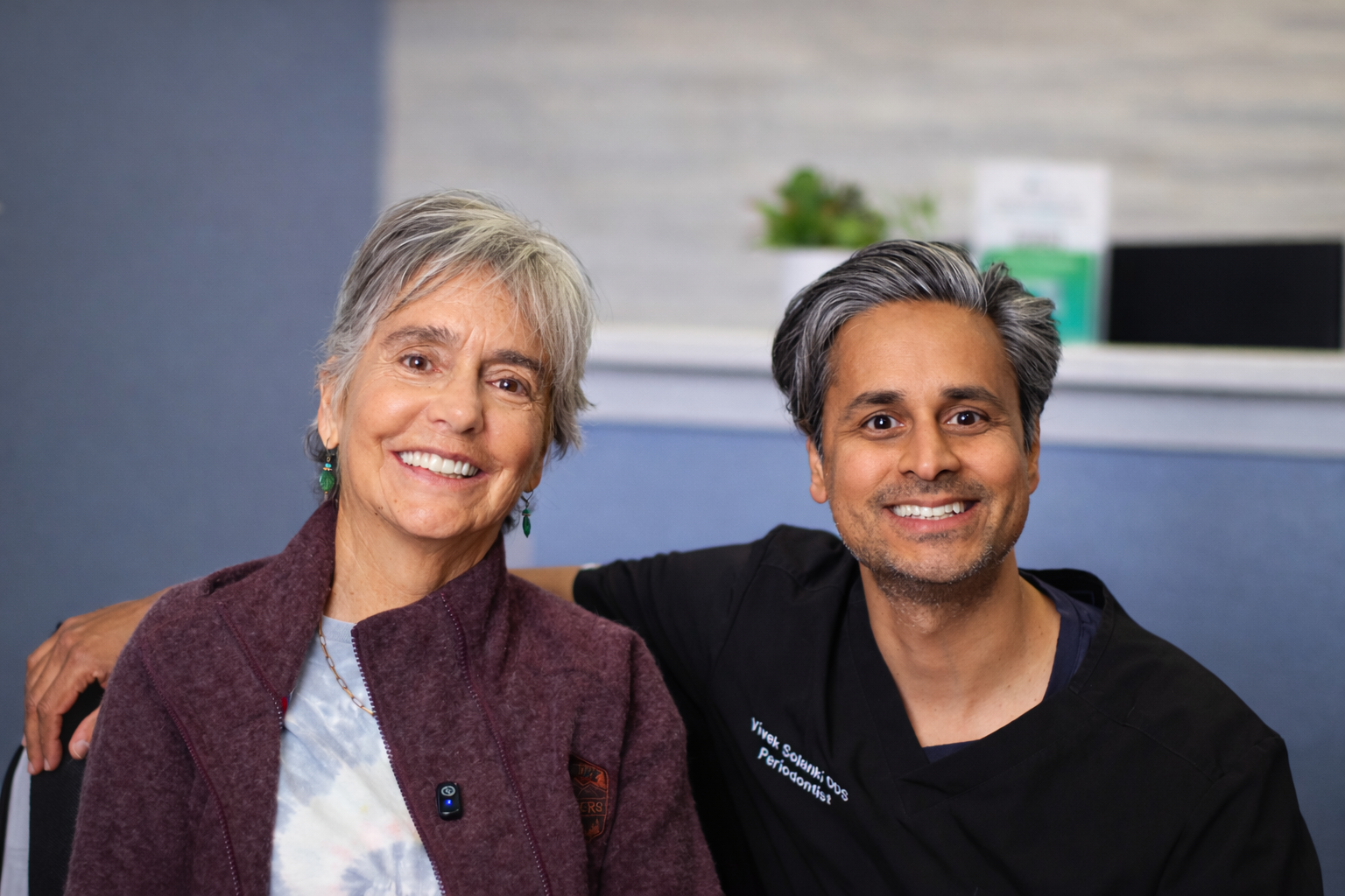 Two smiling people sitting together indoors, one in a purple cardigan and one in a black shirt.