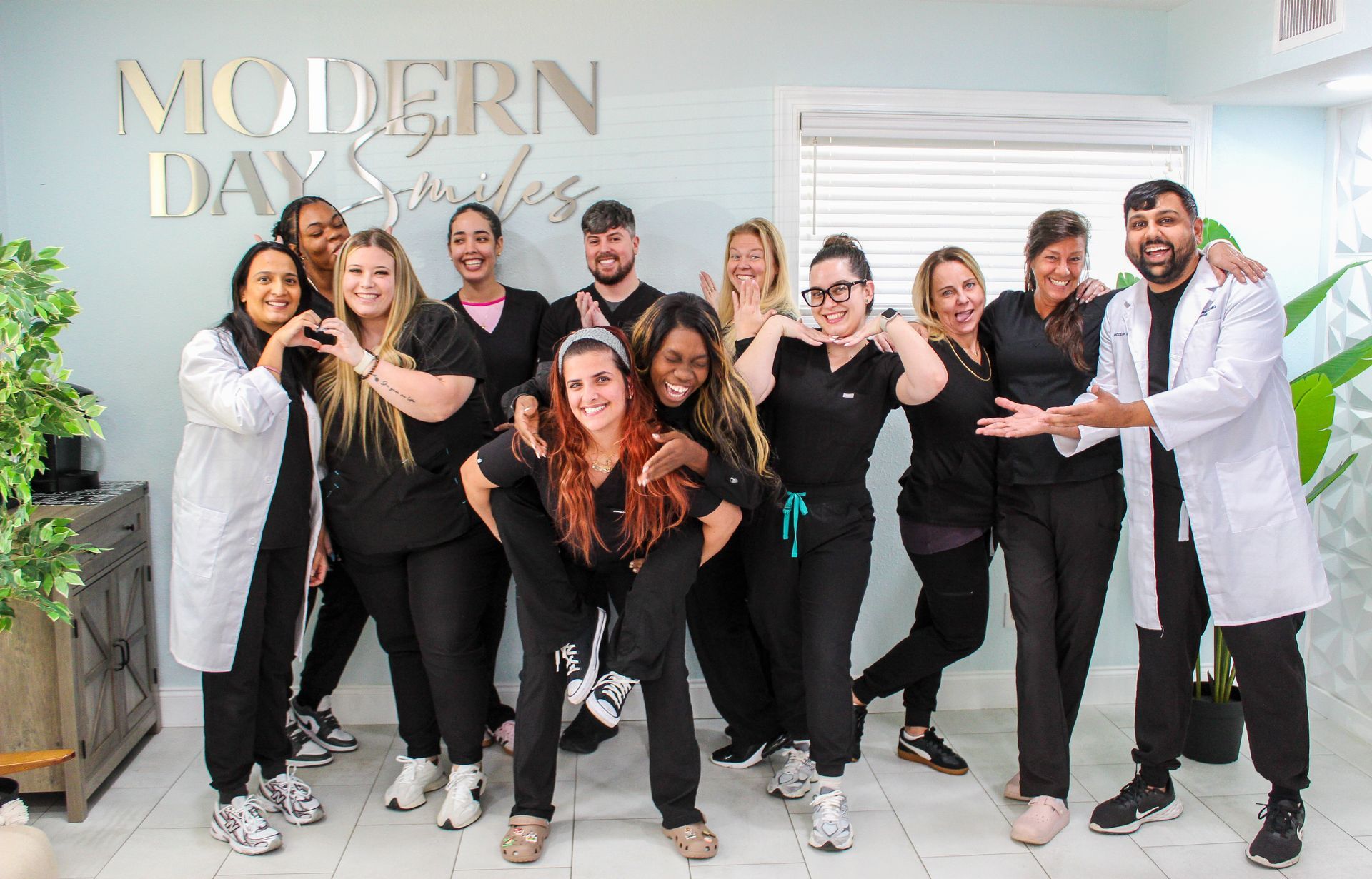Group of dental clinic staff posing playfully. Some wear scrubs, others lab coats. The backdrop is a clinic setting.