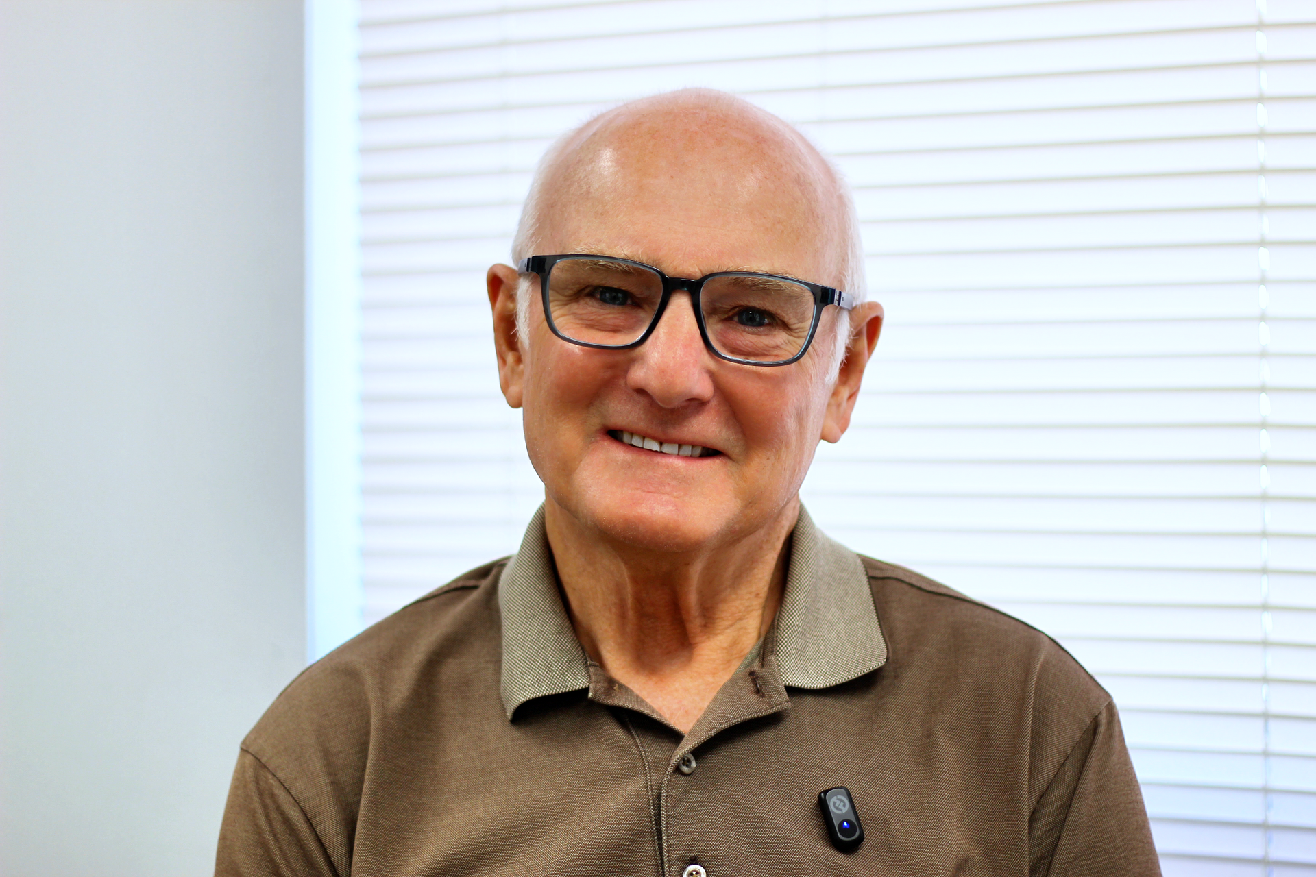 Smiling man with glasses in a brown shirt, seated in front of white window blinds.