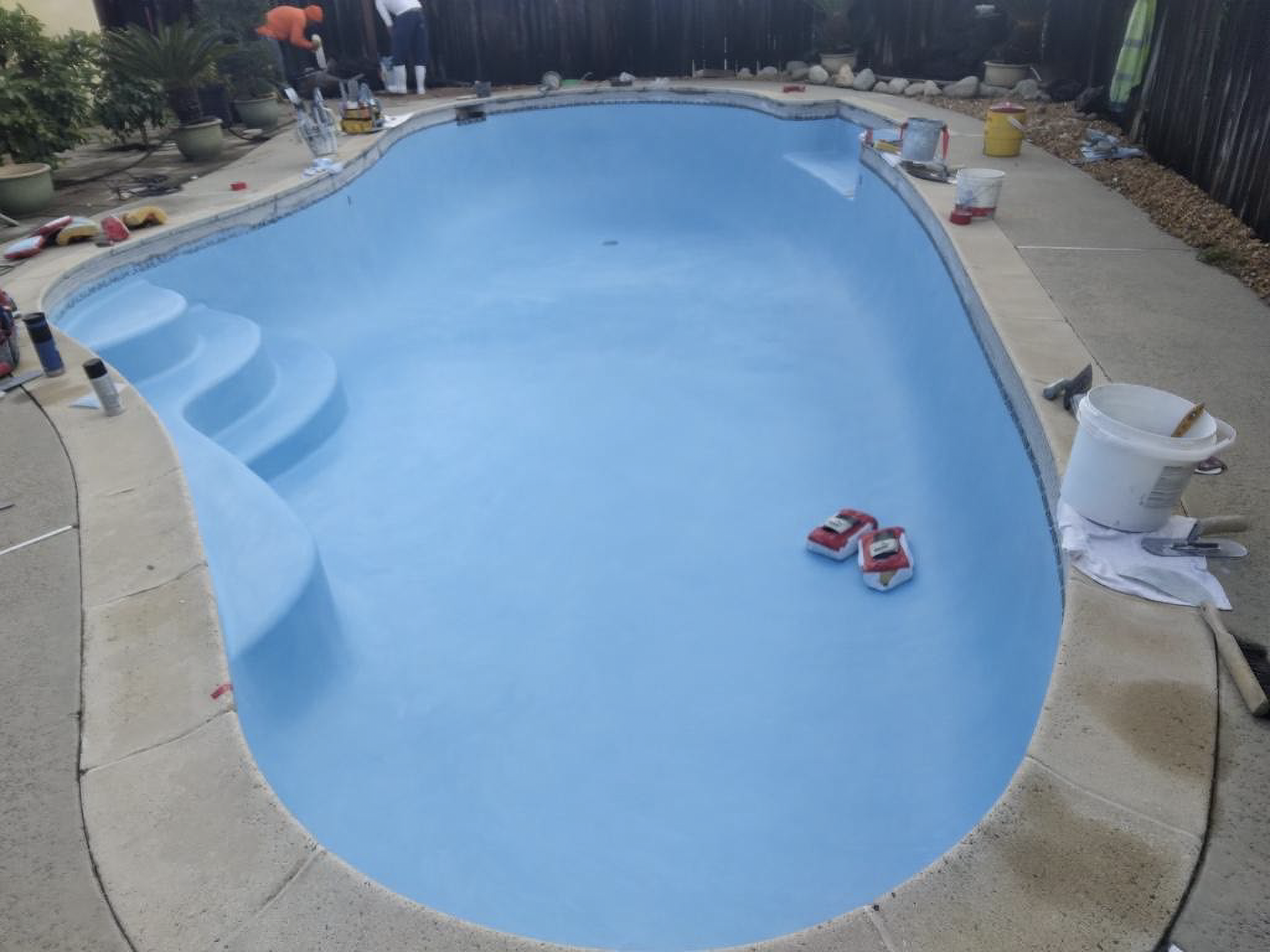 Newly painted light blue swimming pool, steps, surrounded by concrete and tools; people working in the background.