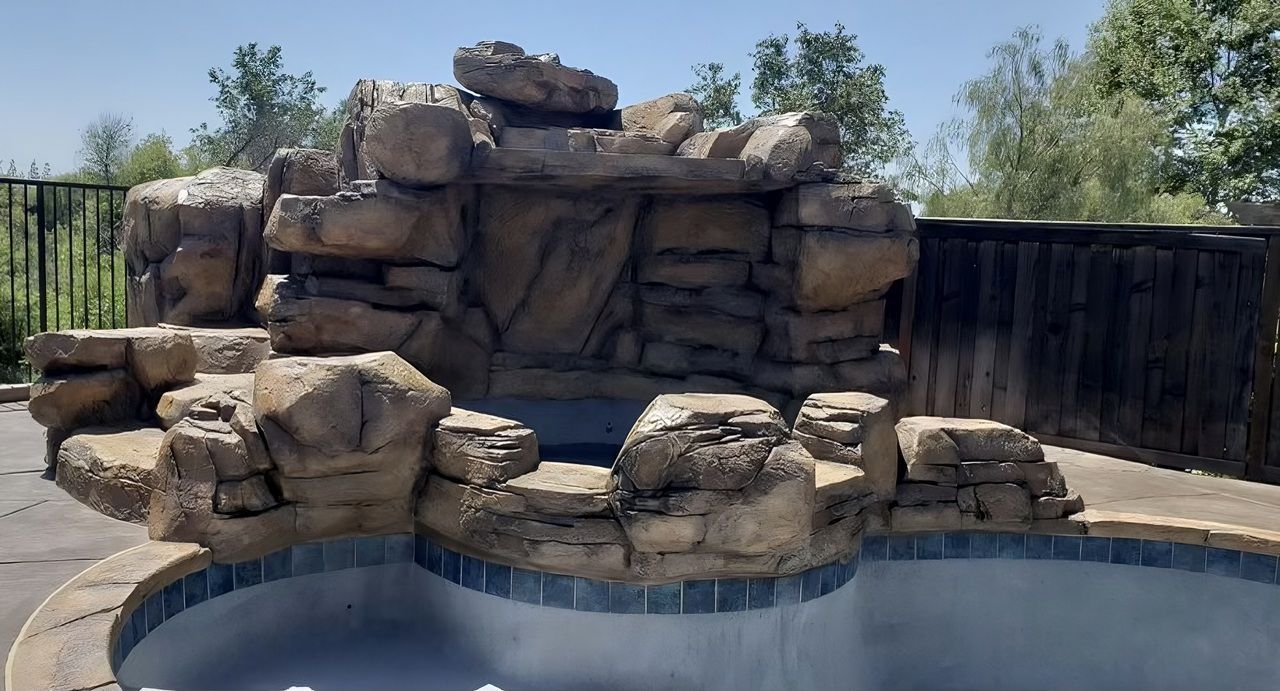 Stone waterfall feature over a swimming pool. Brown rocks and blue tiled edge.