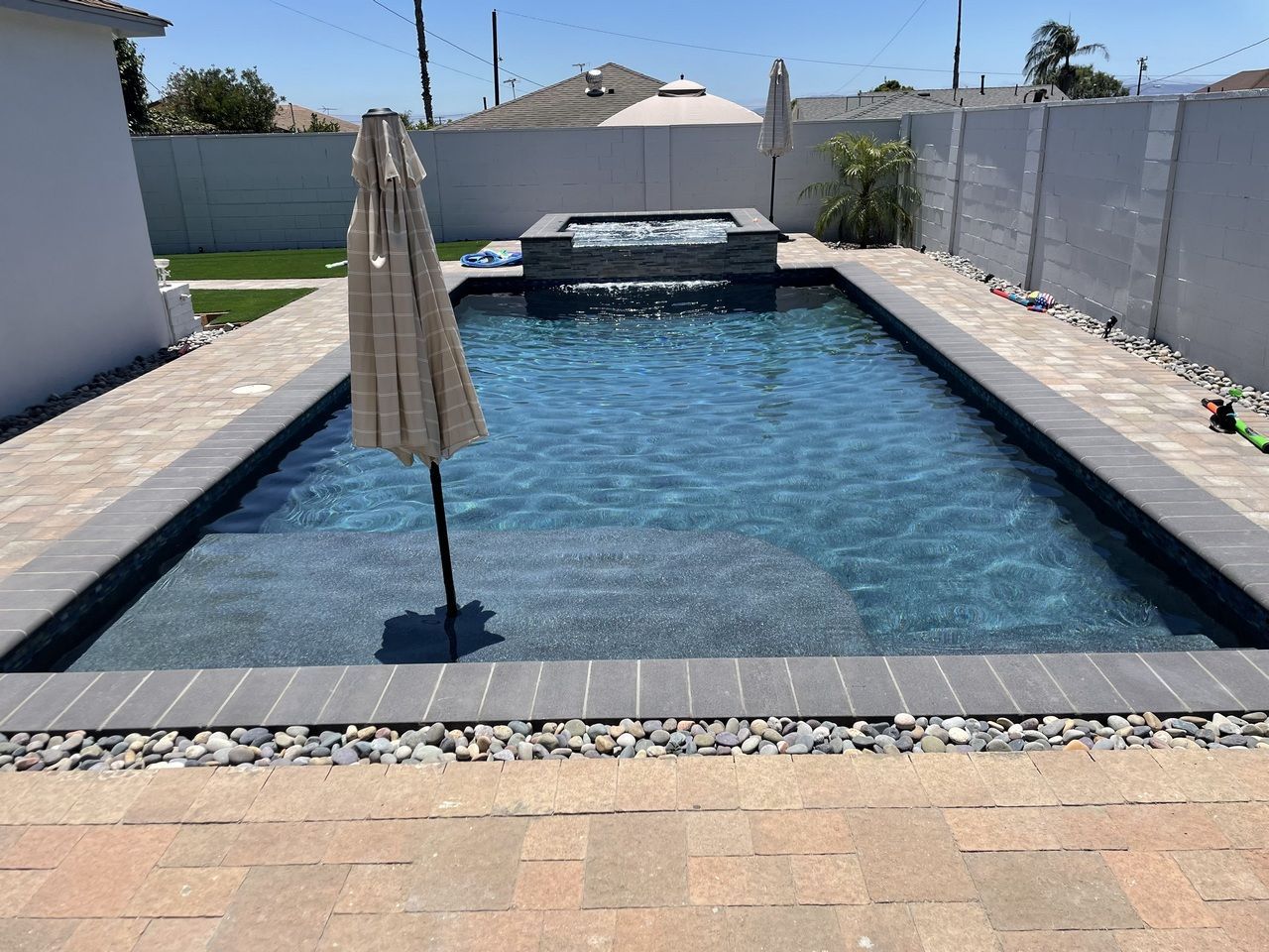Rectangular pool with dark water and a light-colored umbrella in a backyard patio.