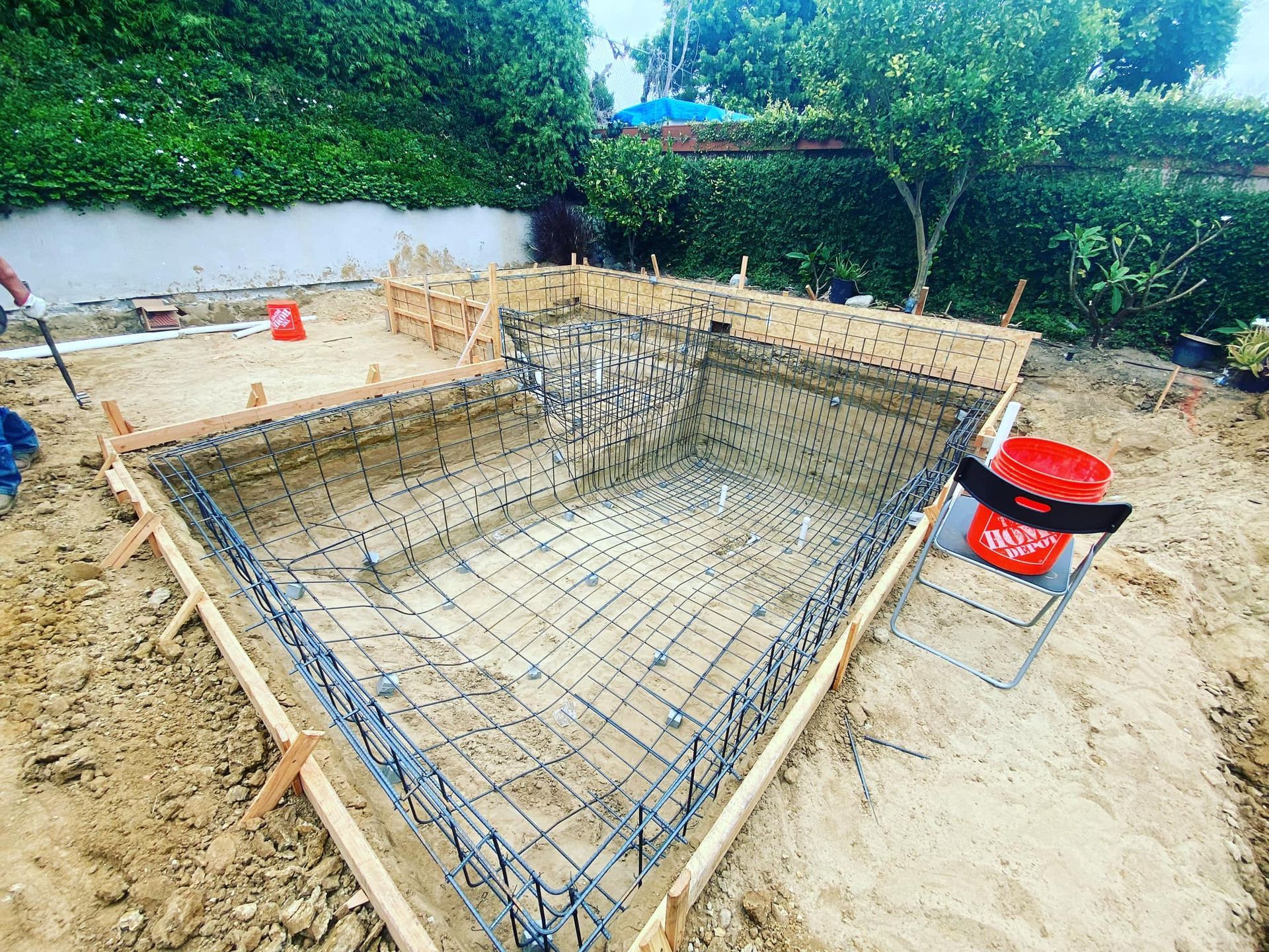 Pool under construction: wooden frame, metal rebar, surrounded by sand, green foliage in the background.