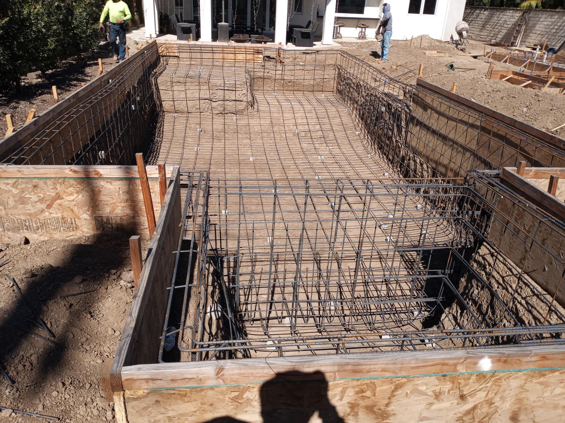 Pool under construction, rebar framework in excavated ground. Two workers near house, wooden forms.