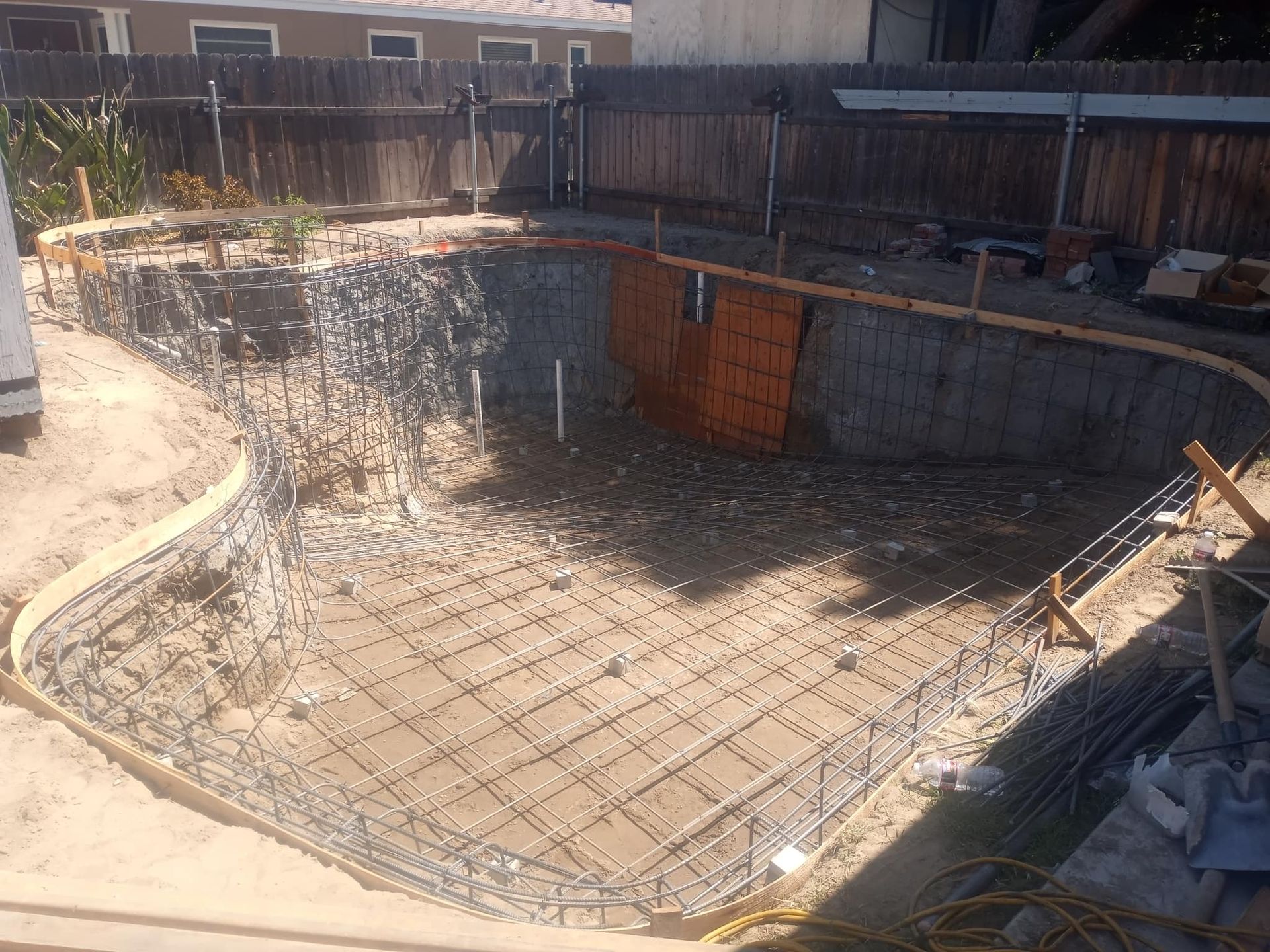 Pool construction: excavated ground, rebar framework, wooden forms for concrete, sunny outdoor setting.