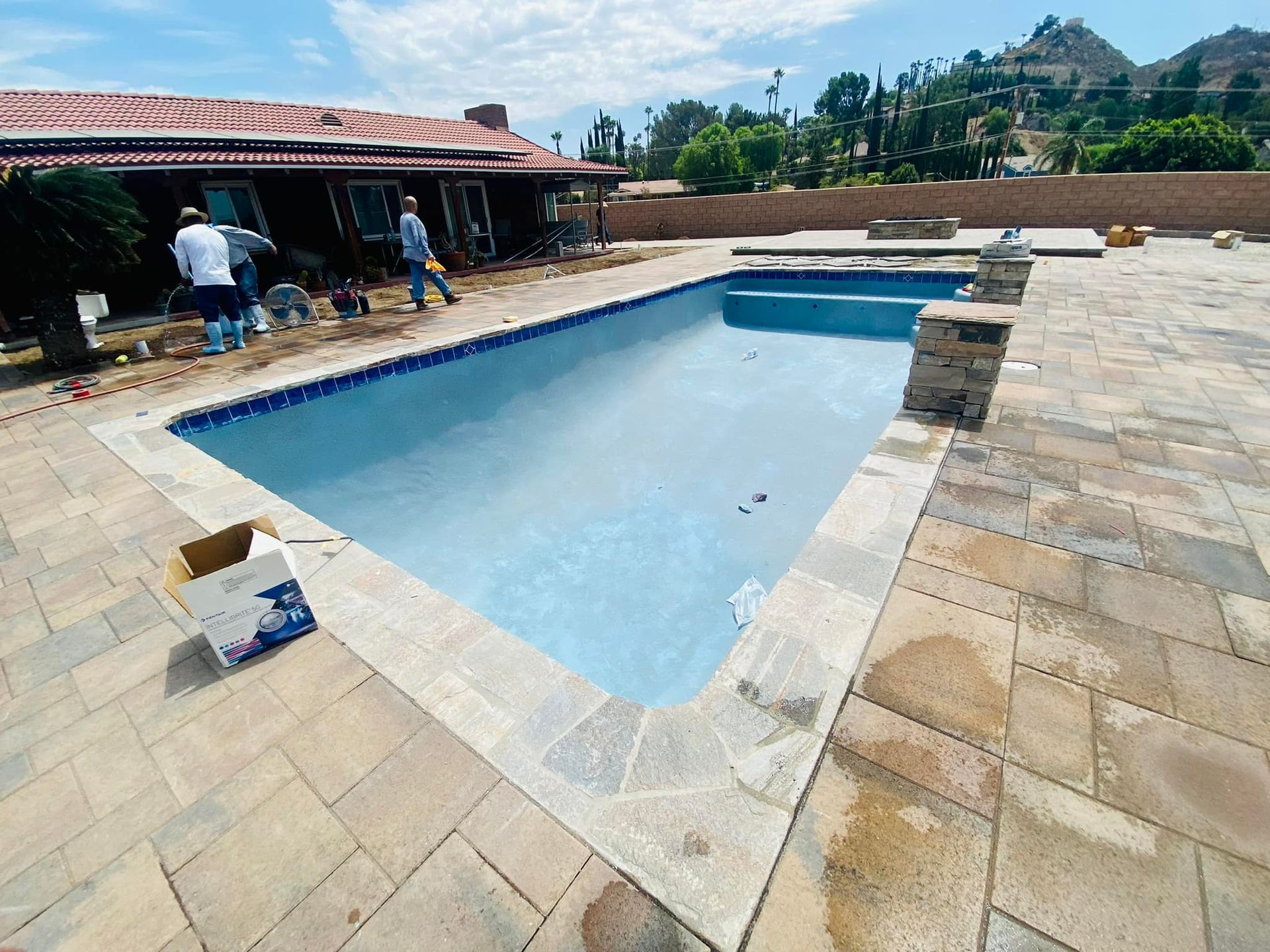 Newly constructed pool with stone coping and brick patio; workers near the house.