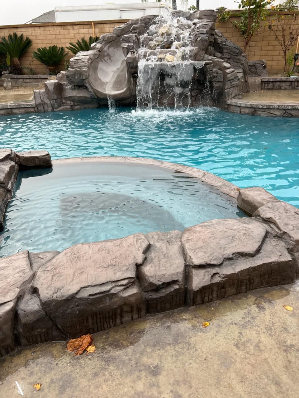 Pool with rock waterfall, slide, and jacuzzi. Blue water, concrete patio, and stone surround.