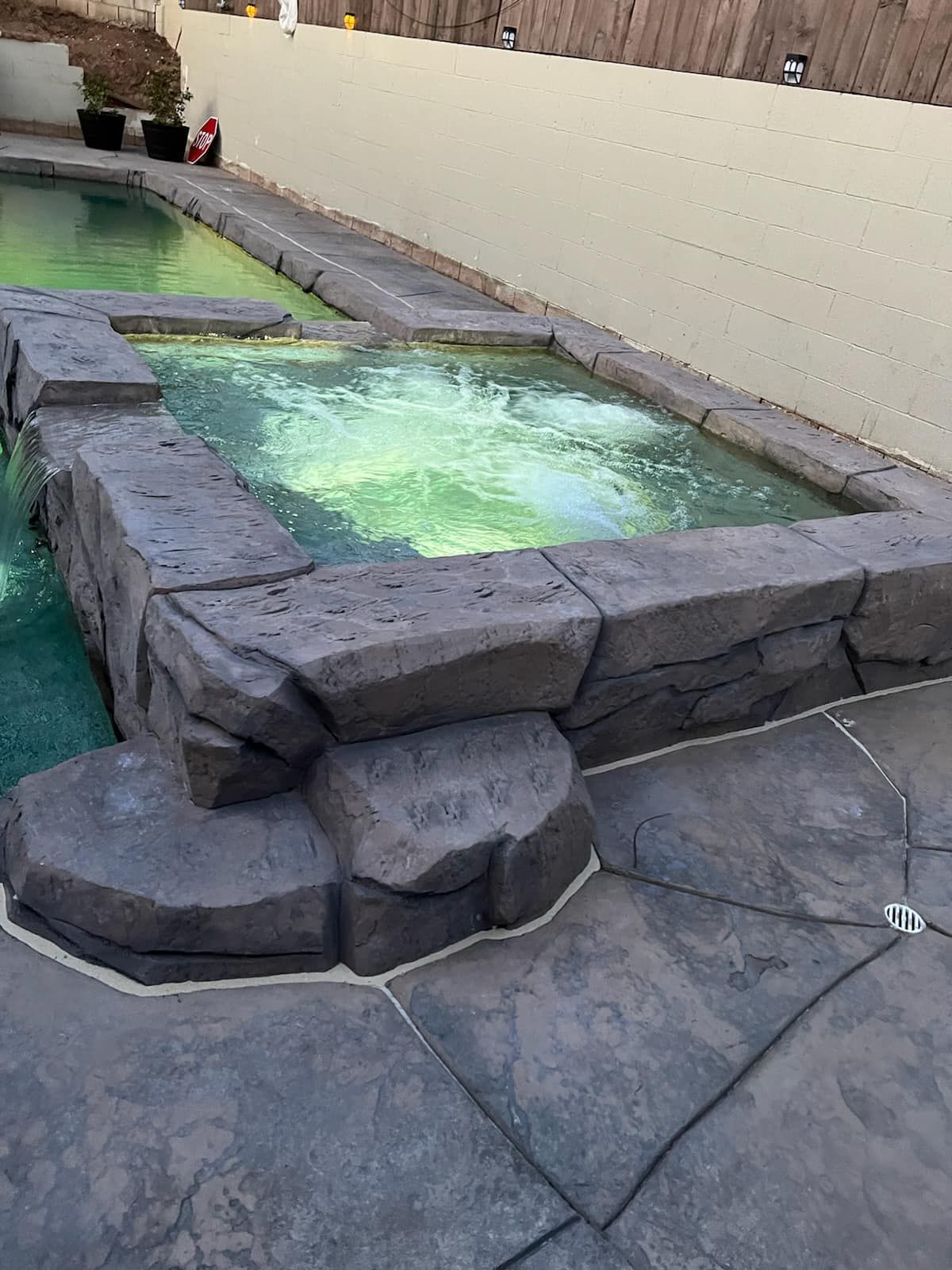 A dark-stone hot tub next to a pool with green-tinted water. Stone patio surrounds them.