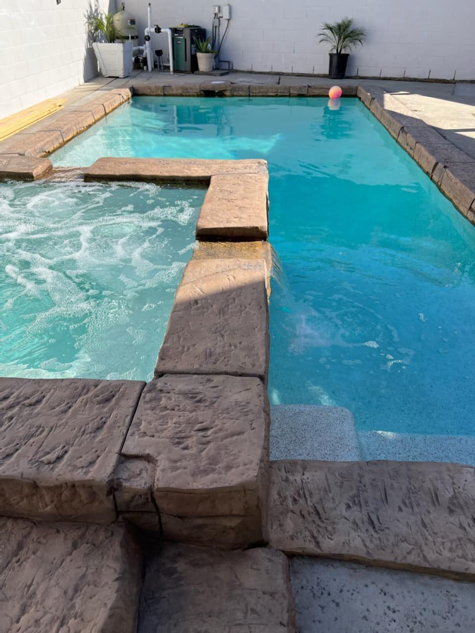 A rectangular swimming pool with stone edging. The water is a bright blue-green, and a pink ball floats in the distance.