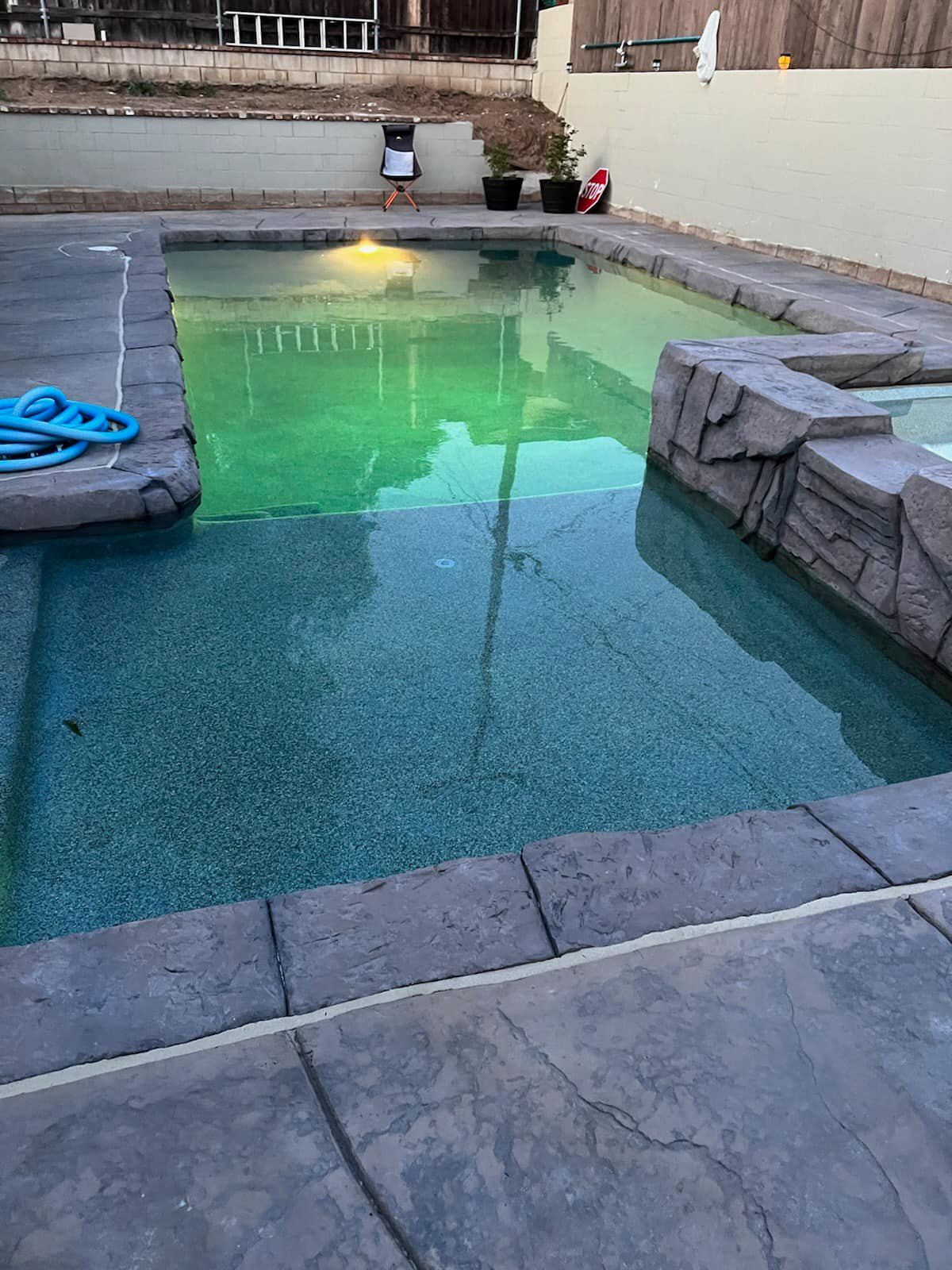 Swimming pool with green tinted water, blue hose, concrete deck, and stone accents.