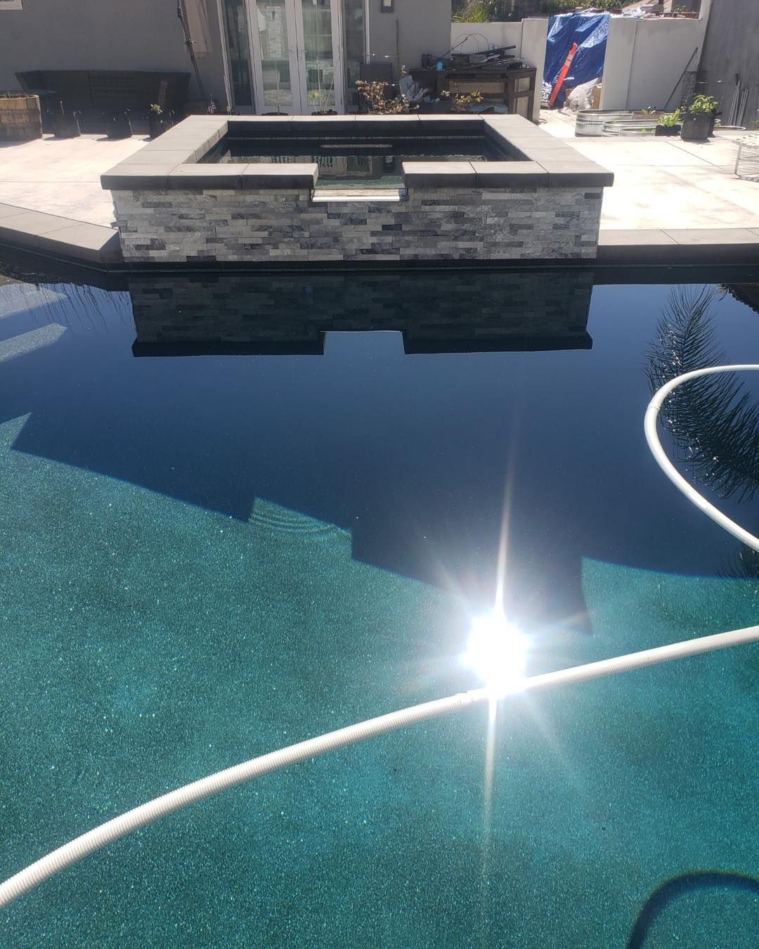 Pool with dark water reflecting a stone structure. The pool bottom is a teal color. A white hose is present.