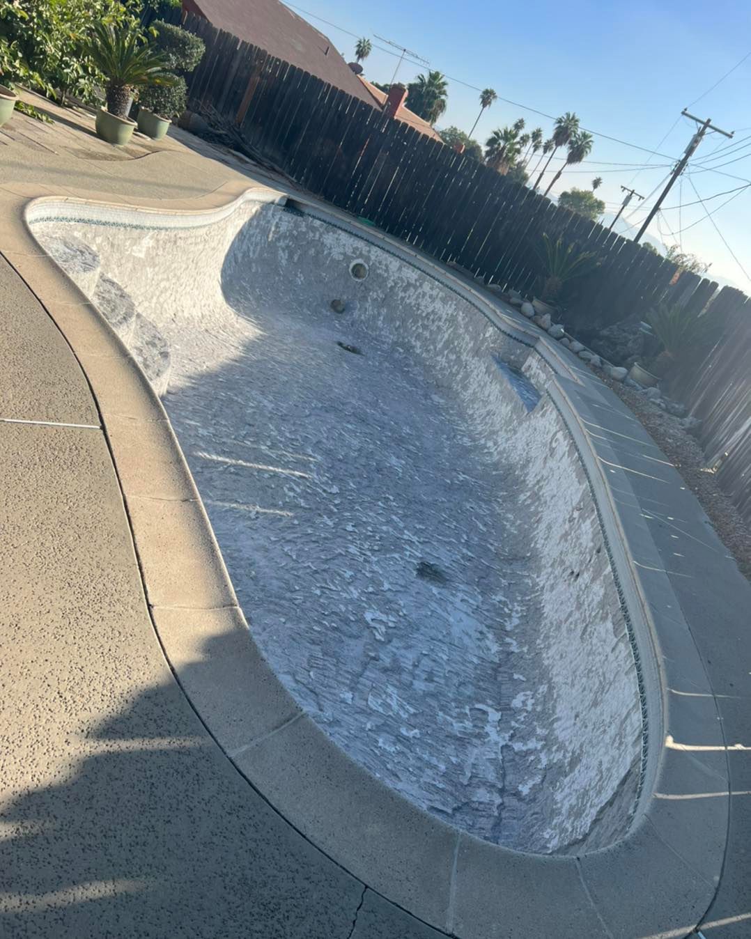 Empty, kidney-shaped swimming pool with concrete surround. Gray with white residue, in a sunny backyard with a fence.