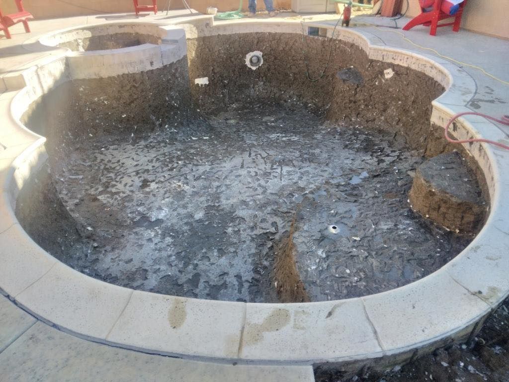 An empty, kidney-shaped pool undergoing renovation, surrounded by concrete. Earth and debris cover the pool floor.