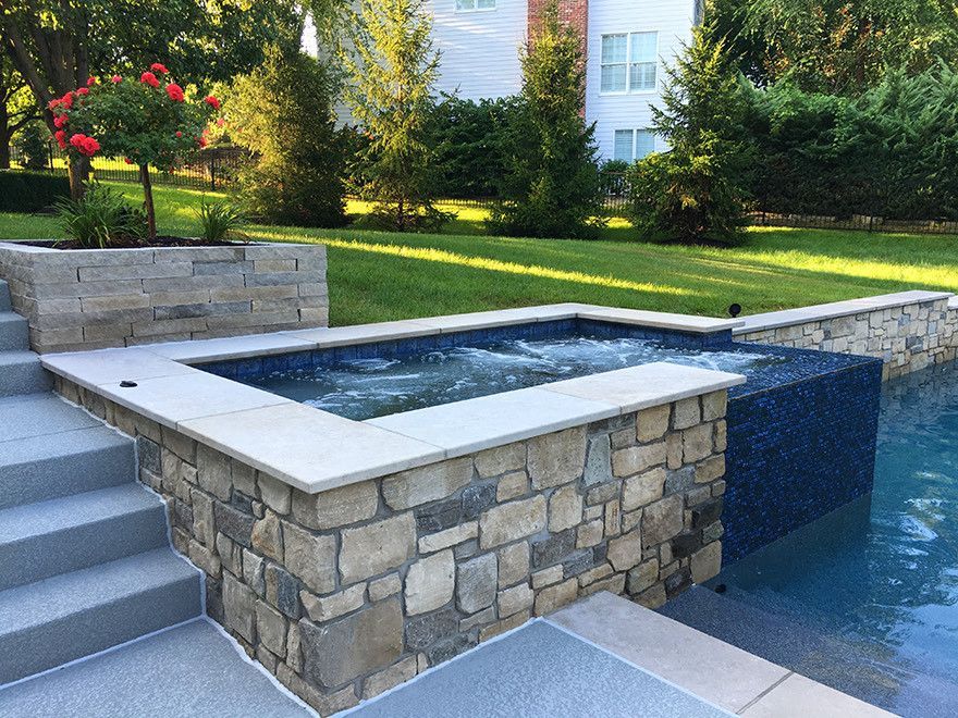 Hot tub next to a pool with stone walls and stairs, set in a backyard with green grass.