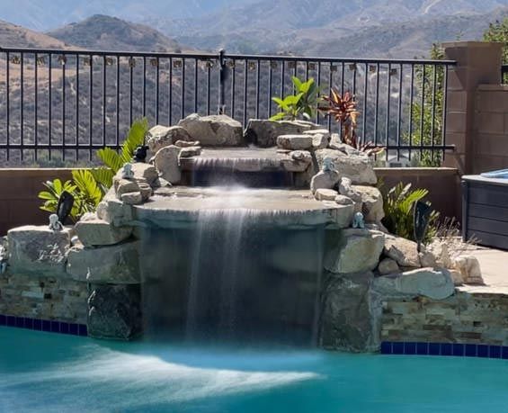 Waterfall feature cascading into a pool, with a mountain backdrop, behind a black metal fence.