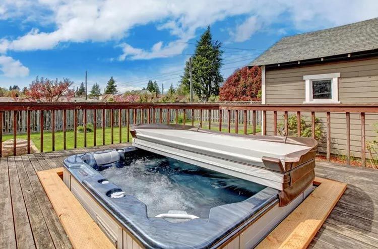 Hot tub on a wooden deck with cover open, next to a brown building, and a green yard with a blue sky.