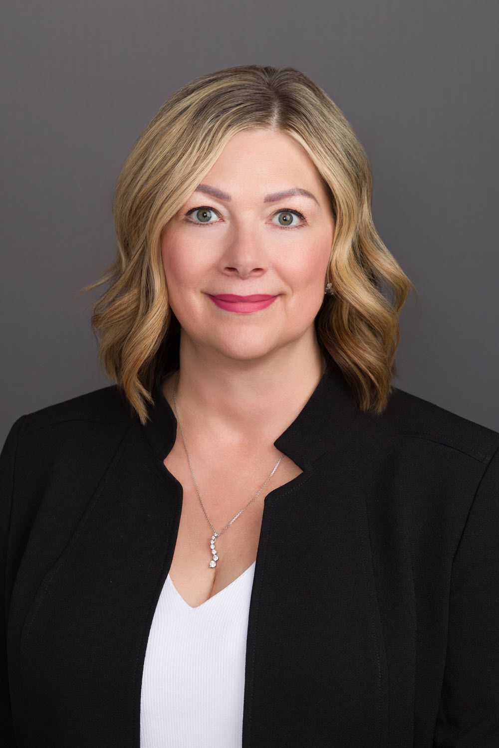 Woman with blonde hair and a black blazer smiles, posed against a gray backdrop.