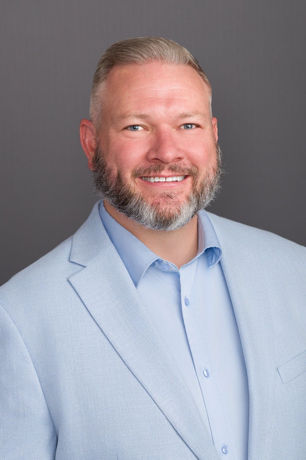 Man with a beard smiling, wearing a light blue blazer and shirt, against a gray backdrop.