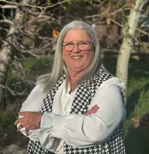 Woman with long silver hair, glasses, arms crossed, wearing a black and white vest, outdoors.