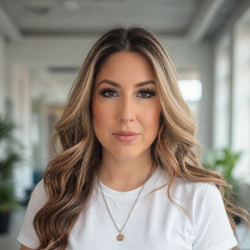 Woman with long wavy brown hair wearing a white shirt, small gold necklace, looking forward in an office setting.