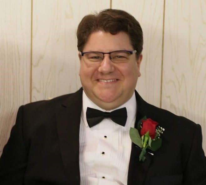 Man in tuxedo with glasses, smiling, wearing a rose boutonniere.