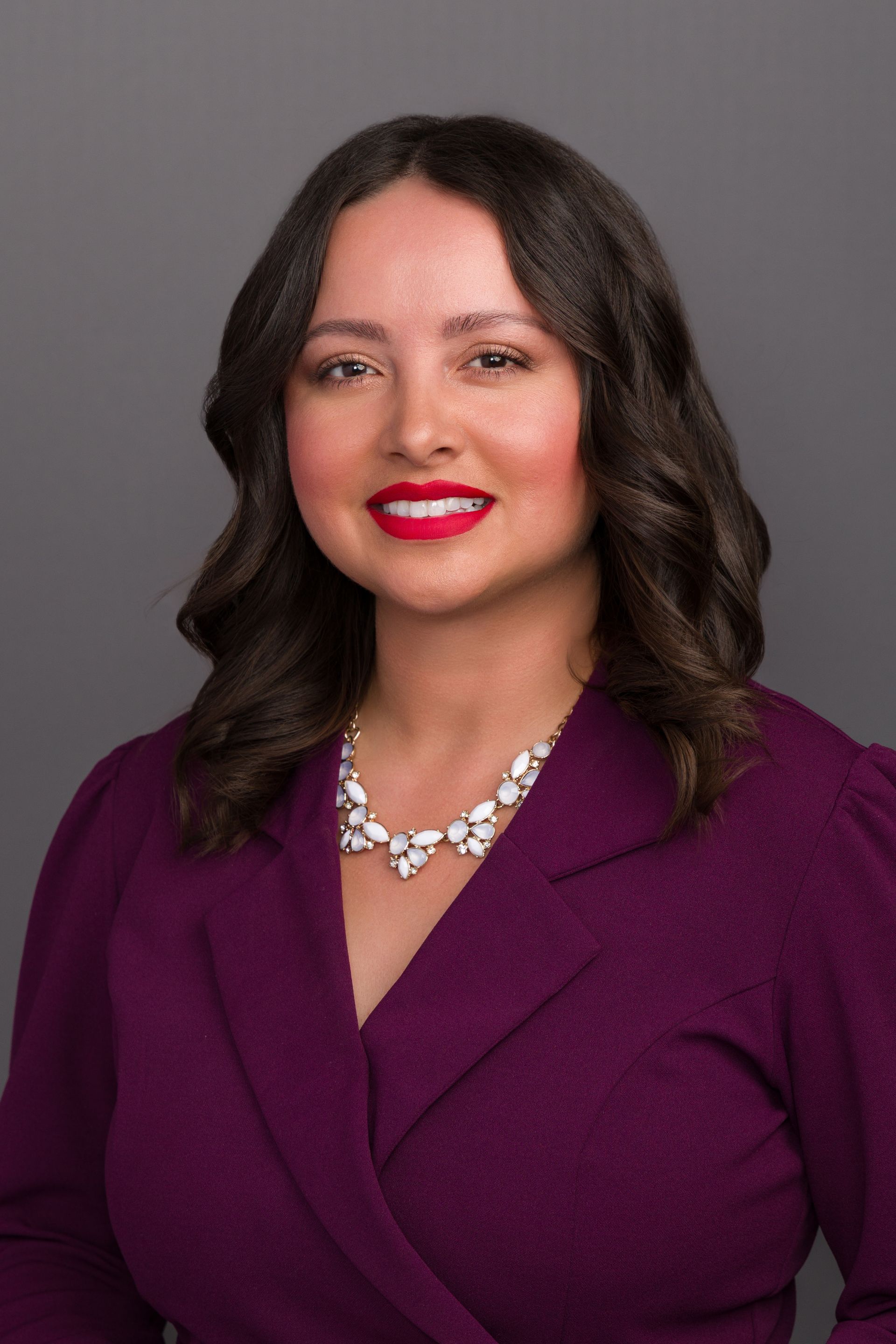 Woman with brown hair, wearing a purple blazer and a sparkly necklace, smiles at the camera.