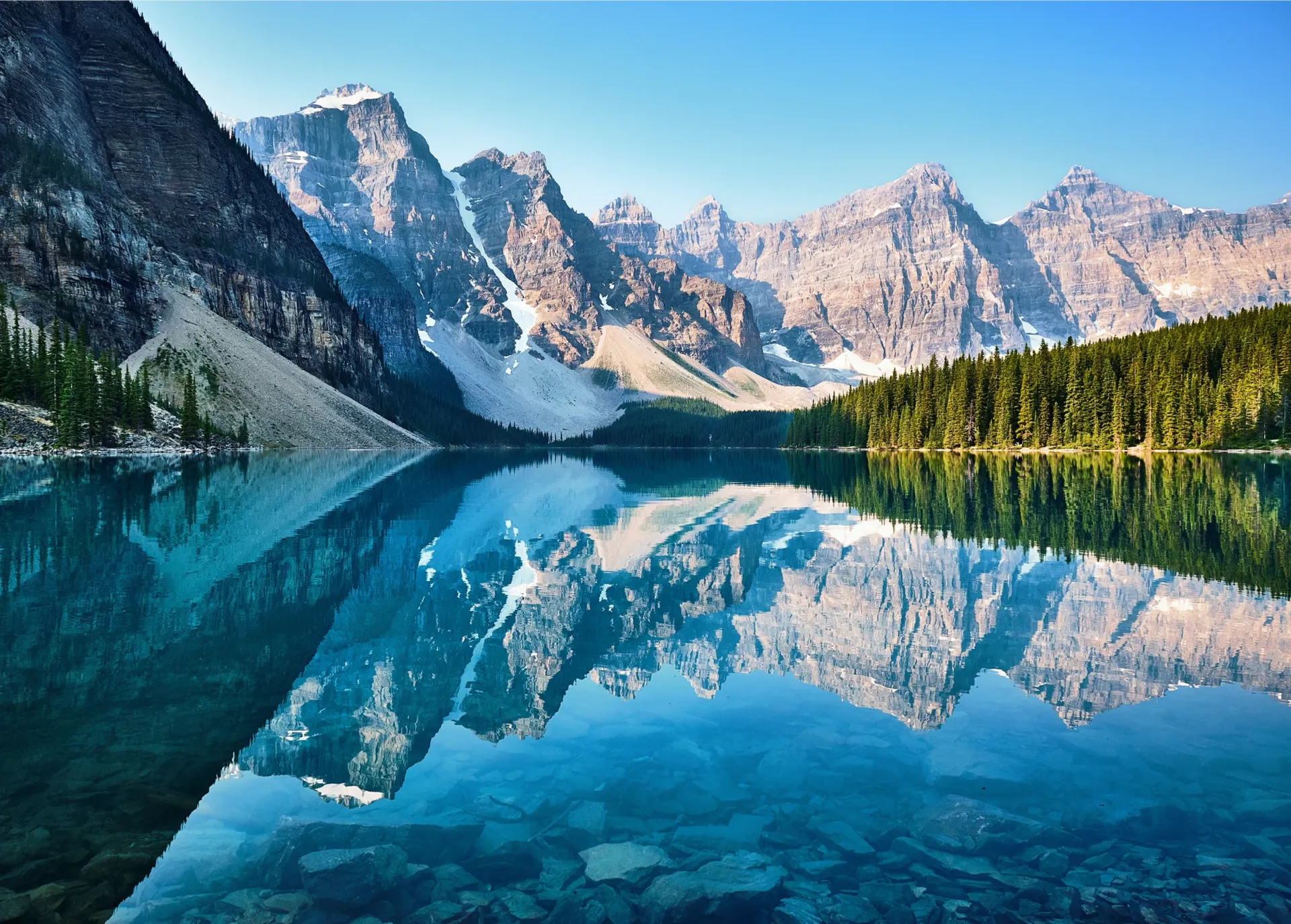 Crystal-clear turquoise lake reflects snow-capped mountains and evergreen trees under a bright blue sky.