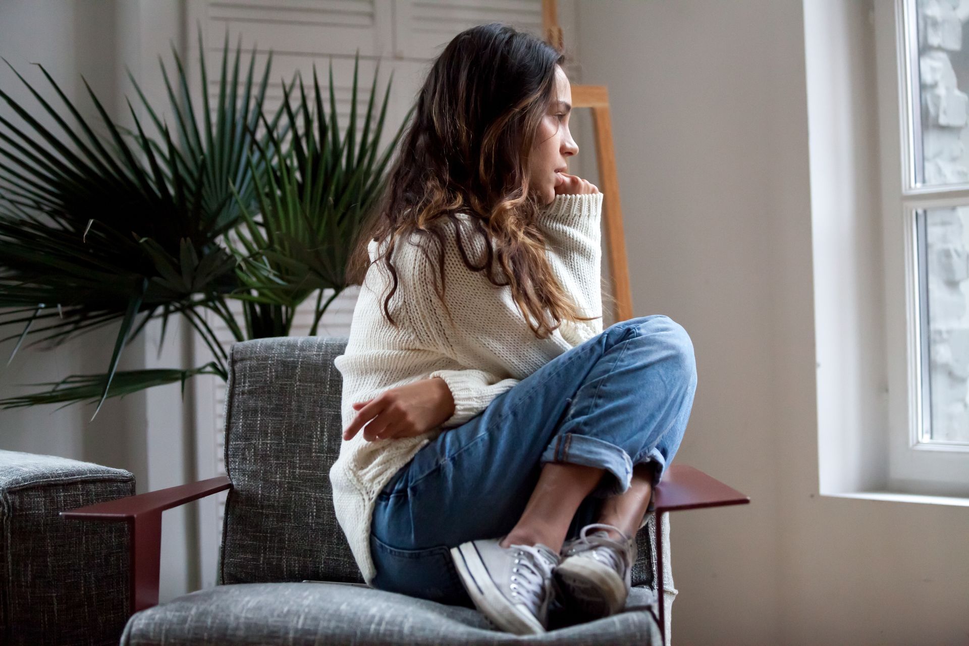 Woman sitting in a chair, looking out the window, wearing a white sweater and jeans.