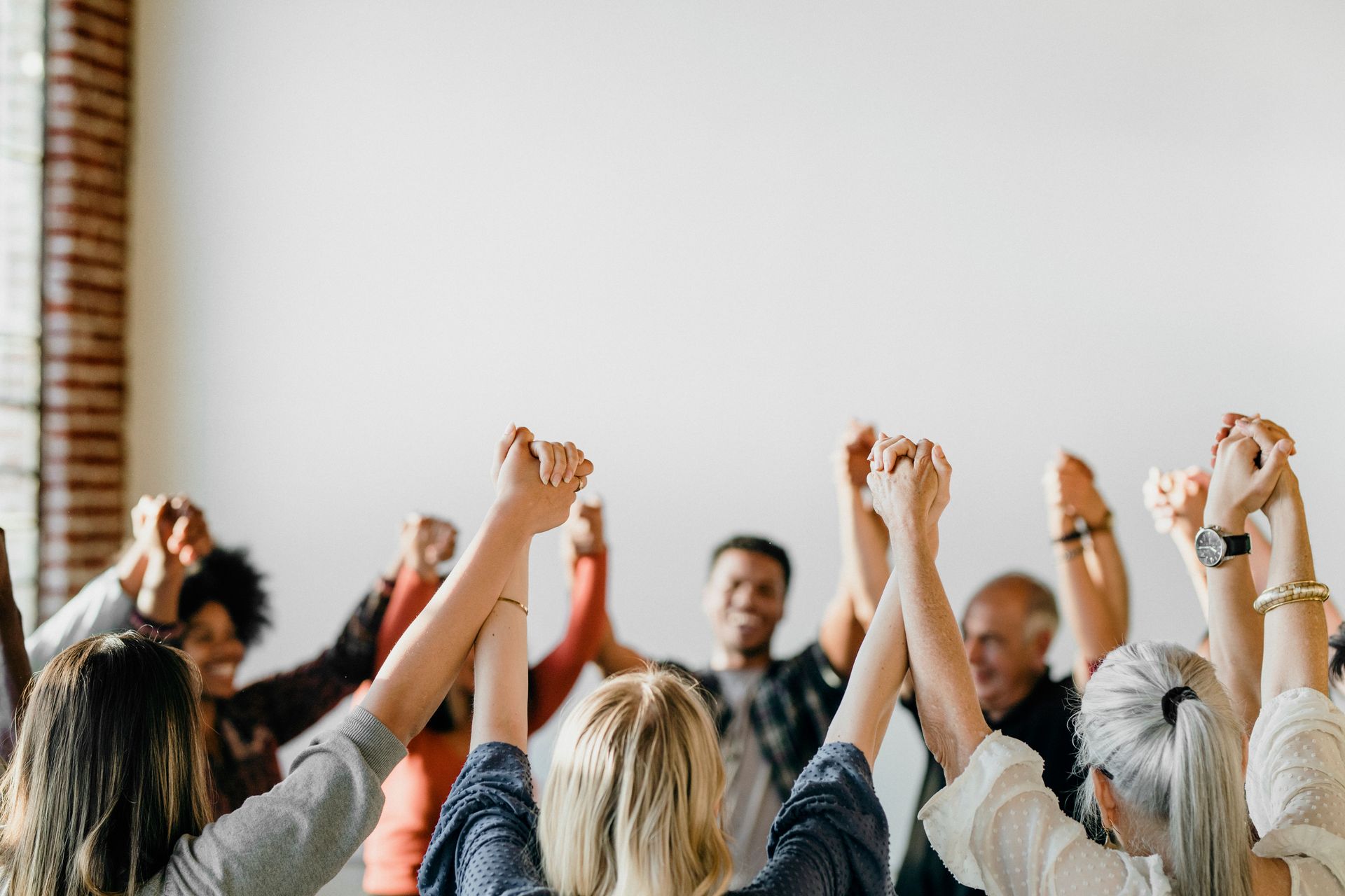 Group of people holding hands up in unity. Inside a white room, arms raised.