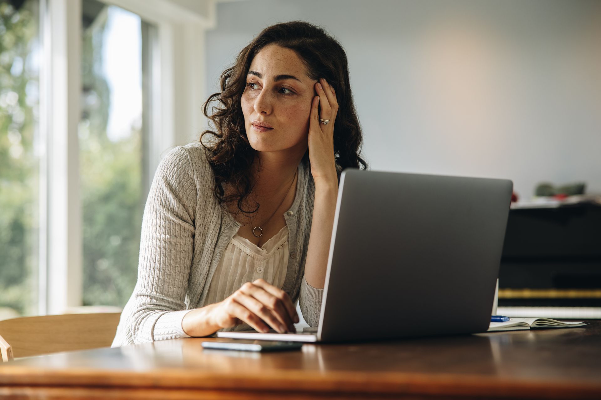 Woman with laptop, looking thoughtful, hand on her head, in a bright room with window.