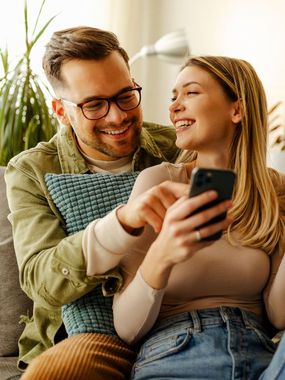 Couple on a couch laughing while looking at a phone. The man points at the screen.