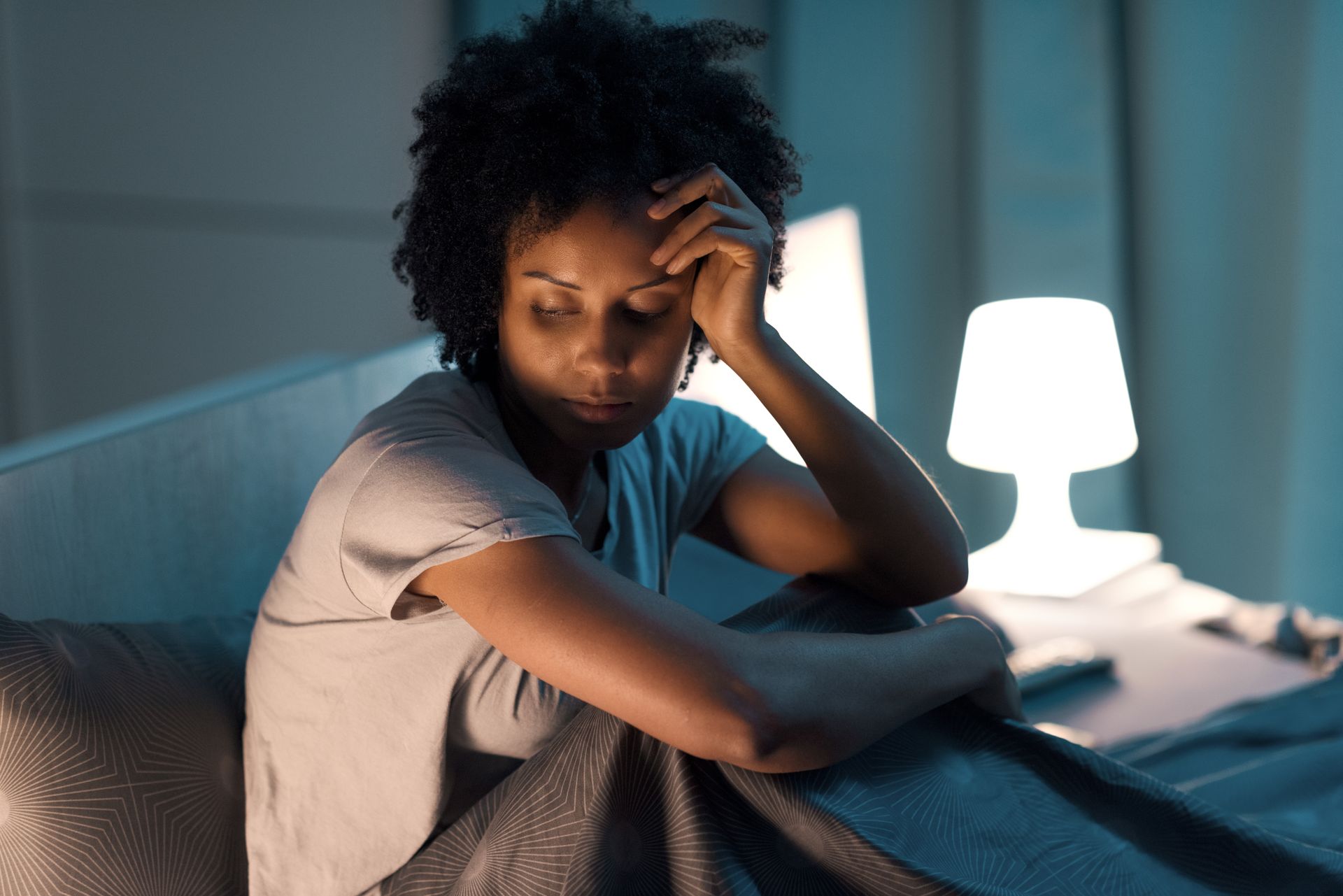 Woman sitting on bed, head in hand, looking down, bedside lamp lit.