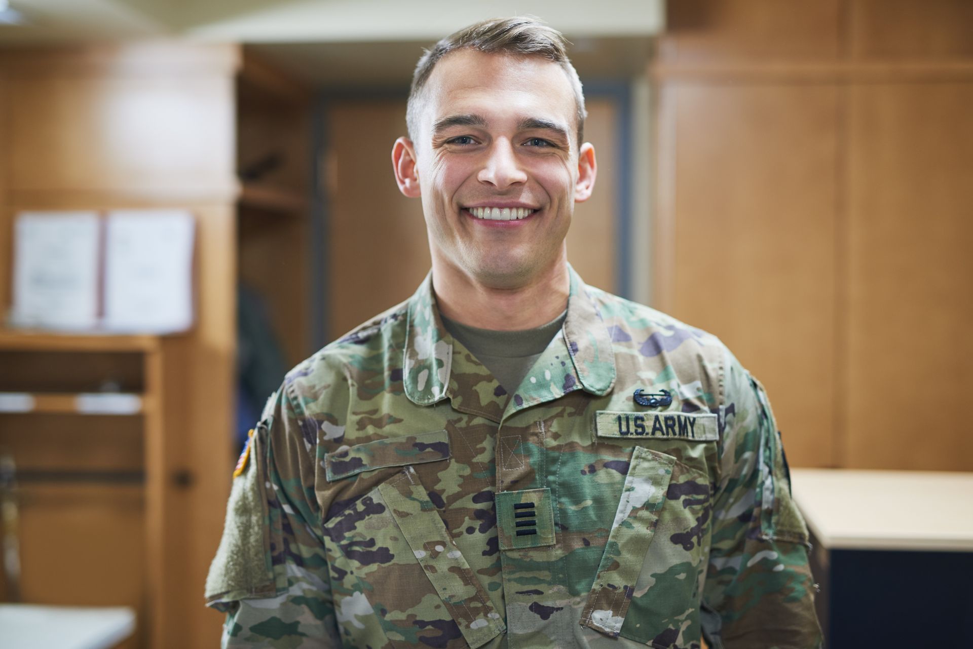 A smiling person in U.S. Army uniform stands indoors.