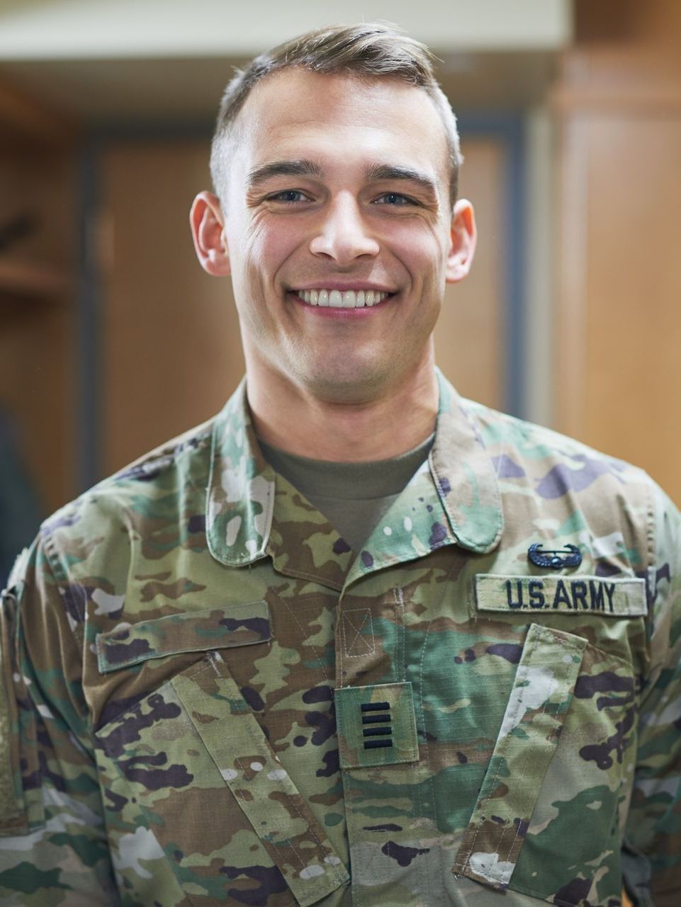 Smiling U.S. Army officer in uniform, name tape and rank visible, indoor setting.
