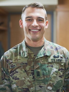 Smiling U.S. Army officer in uniform, name tape and rank visible, indoor setting.