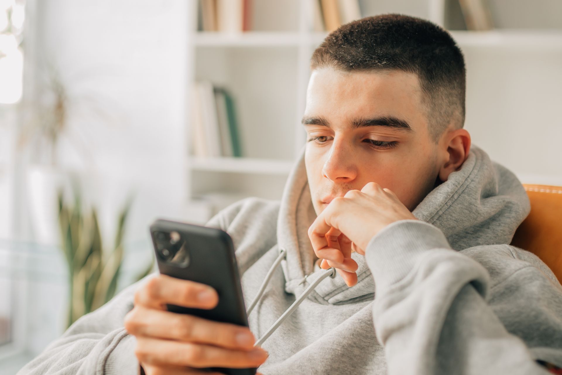 Young person in gray hoodie looking at a smartphone, indoors with a bookshelf.
