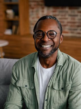 Man wearing glasses, smiling. Light green shirt, white shirt beneath. Brick wall background.