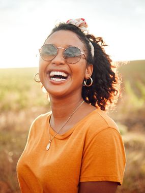 Woman laughing, wearing sunglasses and orange shirt, outdoors in sunlight.