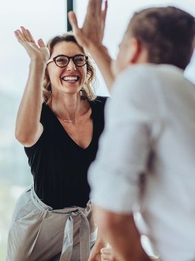 Woman in glasses and man giving each other a high-five, both smiling in an office setting.